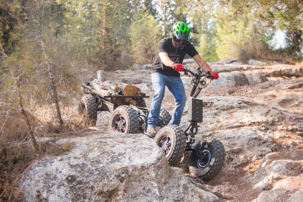 Rider wearing green helmet using an EZRaider HD4 4x4 off-road electric scooter to tow a small trailer loaded with logs over rocky forest terrain